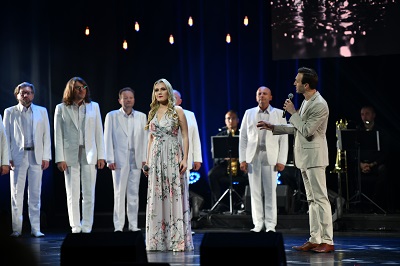 Koncert. Dwoje solistów kobieta i mężczyzna stoją na pierwszym planie za nimi męski chór, wszyscy ubrani w jasne stroje.