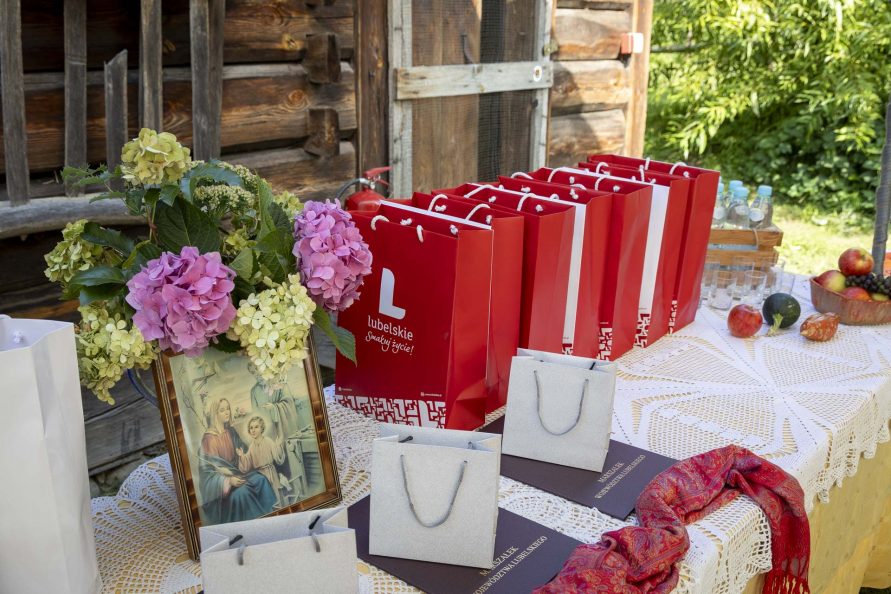 Stół ustawiony na świeżym powietrzu a na nim torebki prezentowe oraz obraz Świętej Rodziny i świeże kwiaty