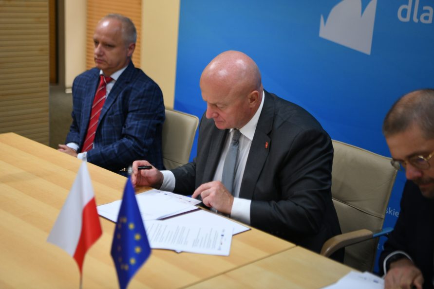 Marszałek Jarosław Stawiarski siedzi przy stole i podpisuje umowę. Po jego Obu stronach siedzą dwaj mężczyźni uczestnicy spotkania