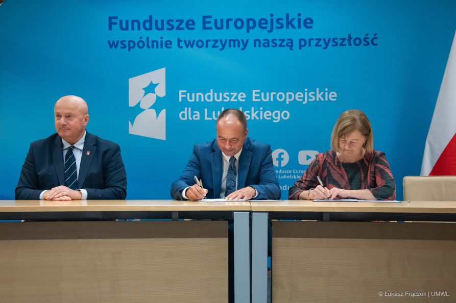 Trzy osoby siedzące przy biurku. Kobieta i mężczyna podpisują dokumenty, obok nich siedzi marszałek Jarosław Stawiarski
