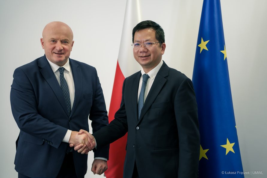 Marszałek Jarosław Stawiarski ściska dłoń Ambasadorwoi Wietnamu na tle flagi Polskiej i Unii Europejskiej.