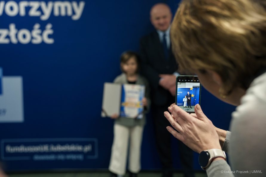 Kobieta robi zdjęcie telefonem marszałkowi i dziewczynce która odebrała nagrodę