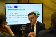 Znajdujący się na sali konferencyjnej uczestnik  spotkania zabiera głos. W tle na ekranie znajduje się informacja, że jest to pierwsze spotkanie partnerów projektu Green Hydra.