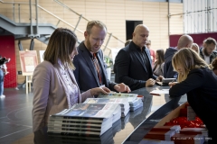 Przy stoisku rejestracyjnym stoi kobieta i dwóch mężczyzn, którzy rozmawiają z rejestratorkami i oglądają wyłożone foldery