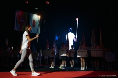 Młoda kobieta przechodzi przez środek sceny i niesie ogień olimpijski