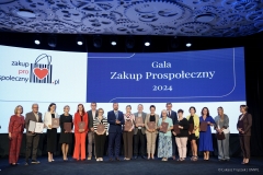 Kilkunastu laureatów certyfikatu stoi na scenie i pozują wspólnie do zdjęcia, za nimi na dużym ekranie  widnieje napis Gala Zakup Prospołeczny 2024