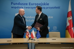 Marszałek Województwa Lubelskiego w geście gratulacyjnym podaje rękę jednemu z beneficjentów programu. Mężczyzna trzyma w drugiej ręce teczkę z dokumentami