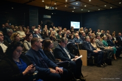 Sala konferencyjna na której znajduje się około stu siedzących osób, uczestników spotkania