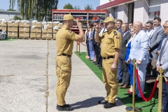 Dwóch strażaków w mundurach salutuje do siebie. W tle widać uczestników wydarzenia około dwudziestu osób. Wszyscy stoją na zewnątrz przed budynkiem w słoneczny letni dzień.