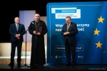 Na scenie znajdują się marszałek i wicemarszałek województwa lubelskiego. Pomiędzy nimi przy mikrofonie stoi ksiądz ewangelicki, który wypowiada się do mikrofonu a w rękach trzyma teczkę z dokumentem dofinansowania