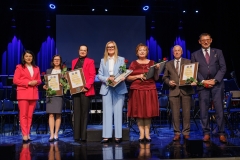 Siedem osób stoi na scenie i pozuje do zdjęć, trzymają w rękach dyplomy i kwiaty