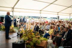 Marszałek Jarosław Stawiarski stoi na scenie i zwraca się do zebranych. Widać około kilkudziesięciu osób siedzących na publiczności. Scena znajduje się pod dużym namiotem i jest ozdobiona bukietami z kwiatów i zbóż